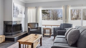 Un salon doté d’une cheminée, de canapés gris, d’une table basse en bois et de grandes fenêtres avec des rideaux.