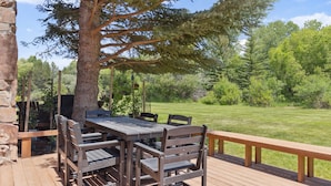 Une terrasse en bois avec une table et des chaises, entourée d’un mur de pierre et offrant une vue sur un champ verdoyant et des arbres.