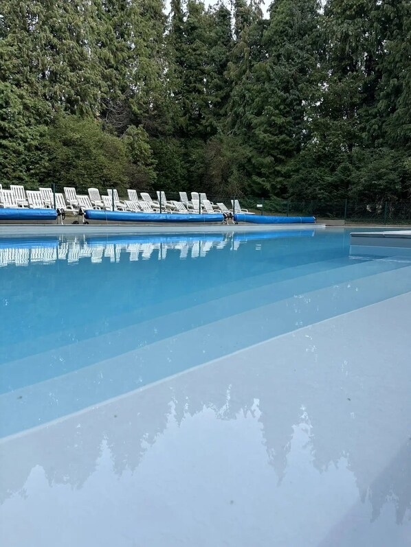 Une piscine à l’eau d’un bleu cristallin, entourée de chaises longues blanches et bordée d’une zone boisée.