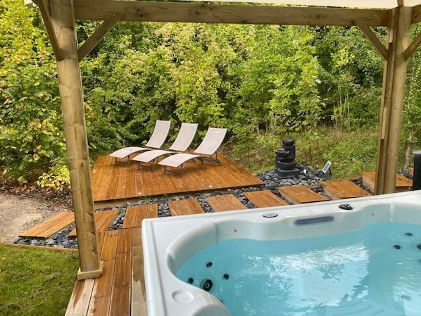A hot tub with a wooden deck and lounge chairs in a garden setting.