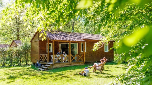 A wooden cabin with a porch, surrounded by greenery. - ZEcamping 4* familial piscine dog friendly locatif TV BBQ 35m2 7 pers (La Bastide-de-Sérou)