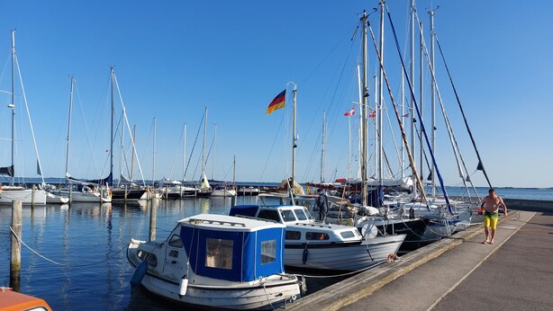 Un puerto deportivo con varias embarcaciones amarradas, cielo despejado y una persona caminando por el muelle.