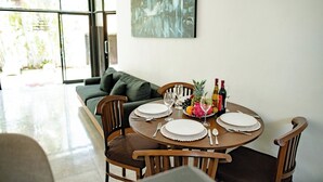 A dining area with a round table set for four, surrounded by wooden chairs. A green sofa is visible in the background, and a large window provides a view of the outdoors. - Saga Villas Oasis - Stylish 2BR villa in excellent location (Bali)