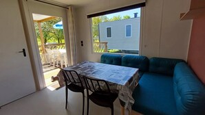 A room with a table covered by a patterned tablecloth, a blue sofa, and a window overlooking a house. - ZEcamping 3 étoiles piscine chauffée chiens acceptés mobil-home BBQ (Lesperon)