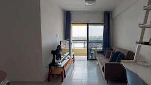 Ein Wohnzimmer mit einer Couch, einem Glastisch, einem Ventilator und einem Laptop auf einem Schreibtisch. Es gibt einen Balkon mit Blick auf Gebäude und einen blauen Vorhang.