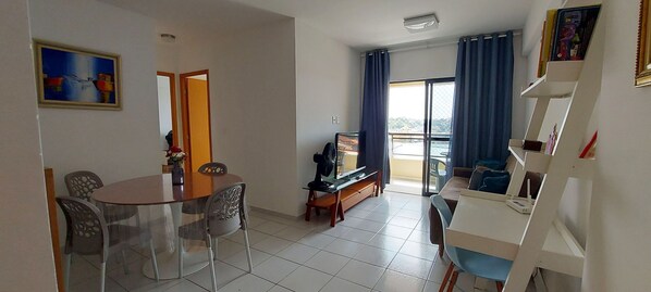 A living room with a dining area, a sofa, a TV, and a balcony with curtains. - COITP0100 - Condomínio Mar de Itapuã (Salvador)