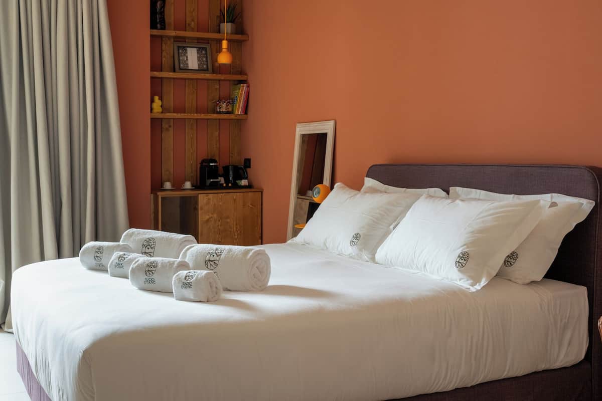 A neatly made bed with white linens and rolled towels, a wooden shelf with books and a coffee maker, and a mirror on the wall.