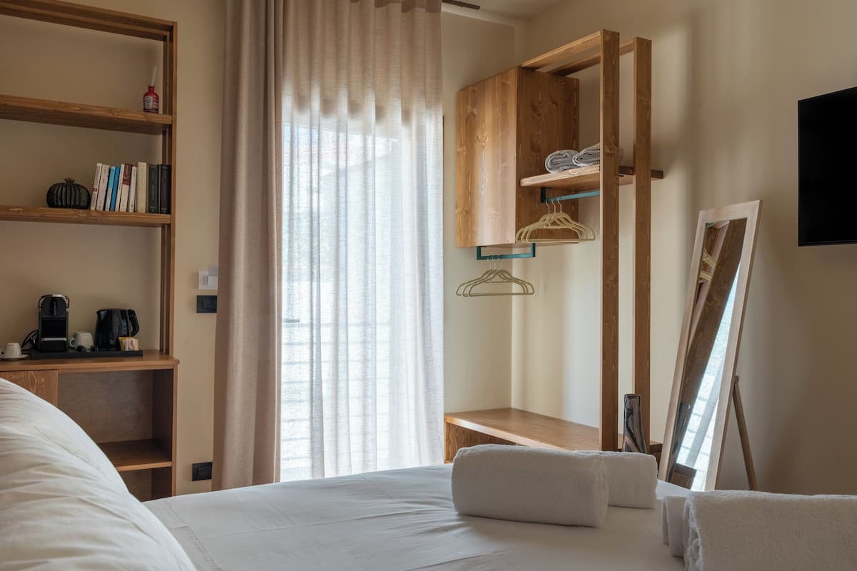 A hotel room with a bed, a wooden bookshelf, a desk with a coffee maker, a mirror, and a window with sheer curtains.