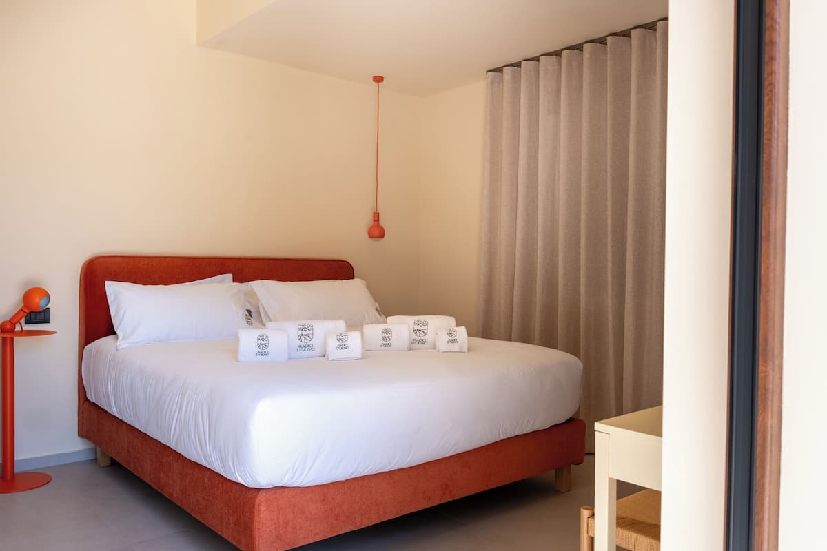 A neatly made bed with white linens and pillows, a red headboard, and a small bedside table with a lamp.