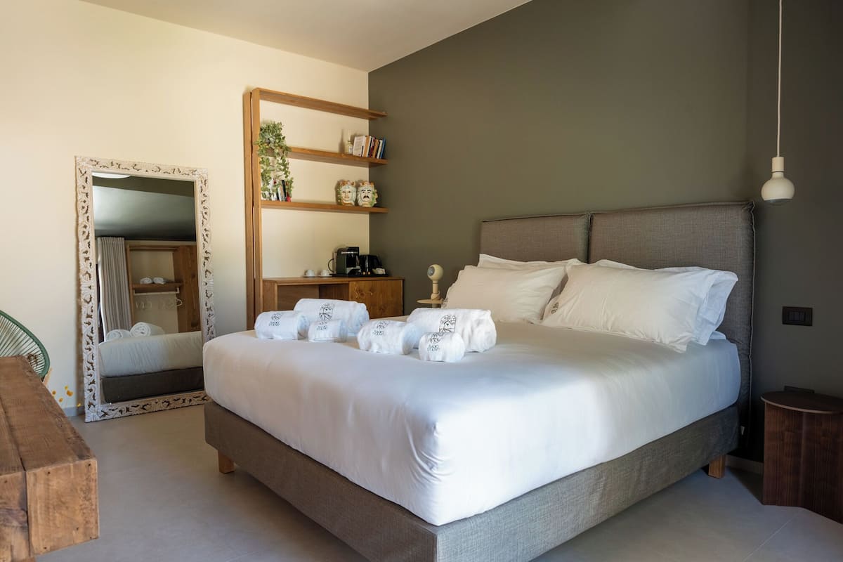 A neatly made bed with white linens and towels, a wooden bedside table, a mirror with a decorative frame, a shelf with books and a plant, and a bedside lamp.