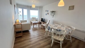 Una sala de estar bien iluminada con un sofá, una mesa de comedor con sillas, un televisor y vista al océano a través de amplios ventanales.