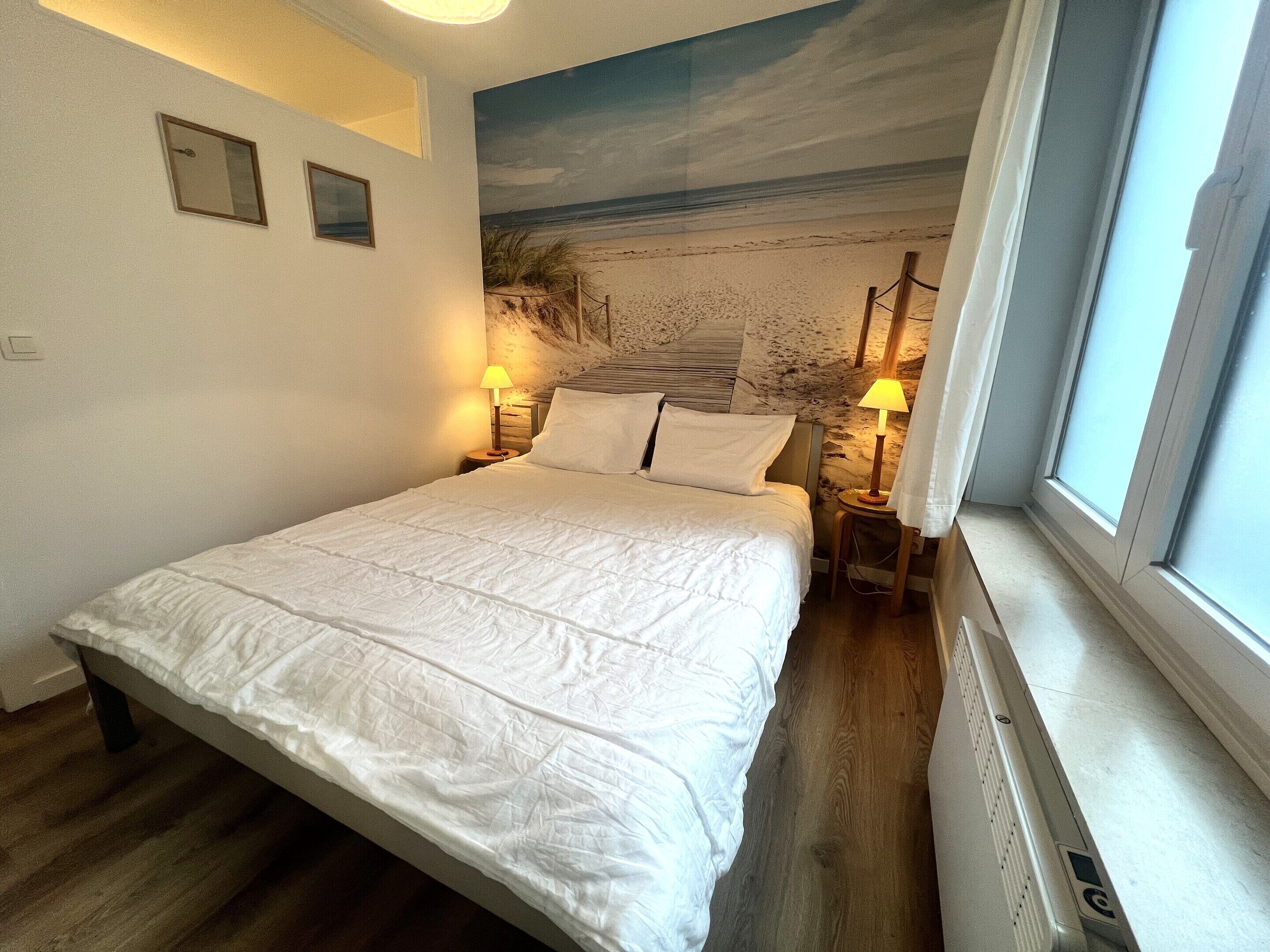 A neatly made bed in a room with a beach mural on the wall, two bedside lamps, and a window with curtains.