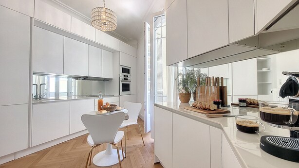 A modern kitchen with white cabinetry, a wooden herringbone floor, a dining area with a white chair, and a hanging light fixture.