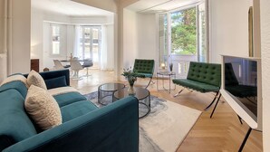 A spacious living room with a large TV, two green armchairs, a blue sofa, and a chandelier.