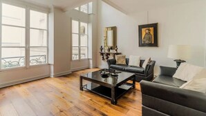 A spacious living room with a black leather sofa, a glass coffee table, and a large window providing natural light. - Magnificent duplex in the Marais district (Paris)