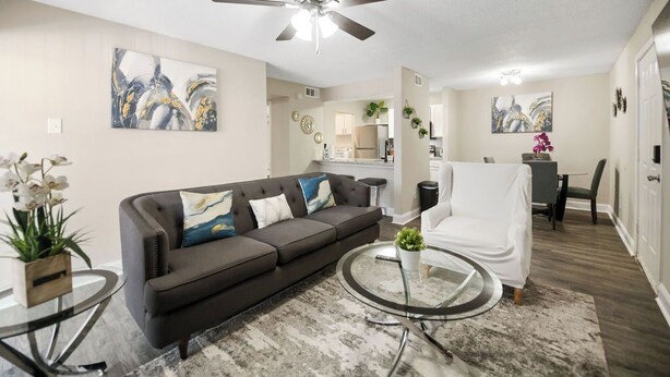 A modern living room with a dark brown sofa, a round glass coffee table, and a ceiling fan. There is a kitchen in the background with white cabinets and a dining area. - I014 - Upgraded 2BD | Pool | DFW South 183/161 (Irving)