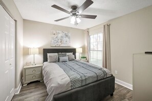 Ein Schlafzimmer mit Bett, Nachttischen, Deckenventilator, Fenster mit Vorhängen und Wandbild.