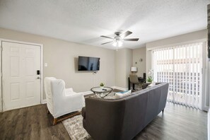Ein Wohnzimmer mit grauem Sofa, einem weißen Sessel, Deckenventilator, Flachbildfernseher und einer Glastür, die auf einen Balkon führt.