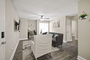 A living room with a ceiling fan, a gray sofa, a white chair, and a gray rug. - I014 - Upgraded 2BD | Pool | DFW South 183/161 (Irving)