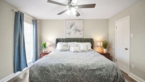 A bedroom with a bed, a ceiling fan, a window with curtains, and a door. - I014 - Upgraded 2BD | Pool | DFW South 183/161 (Irving)