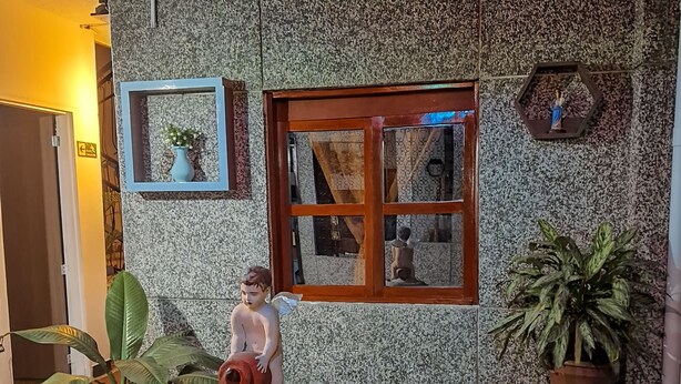 Una fuente de piedra con una estatua infantil, rodeada de plantas en macetas y una pared con azulejos, una ventana y un cuadro enmarcado.