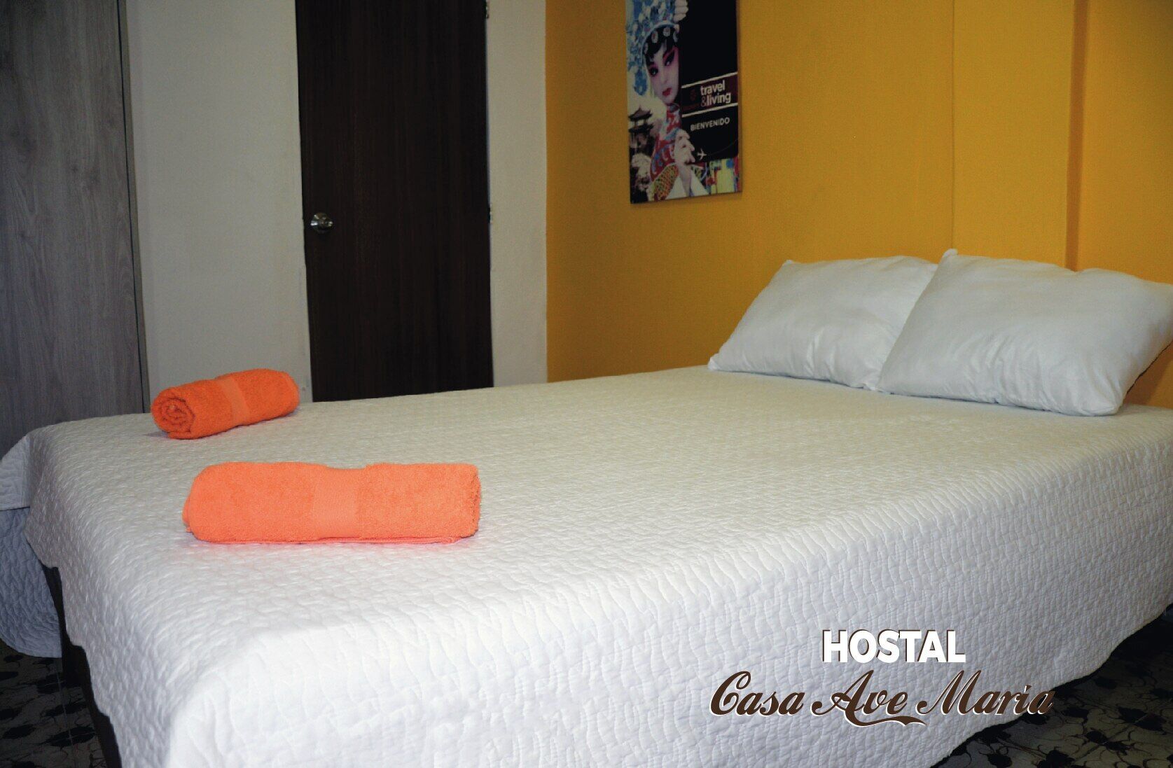 Una cama bien tendida con ropa blanca y dos toallas naranjas, un cartel en la pared y una puerta al fondo.