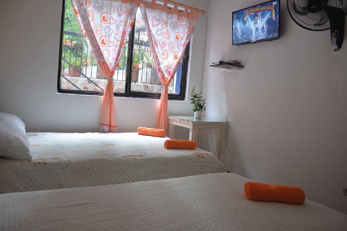 A bedroom with two beds, a window with curtains, a TV on the wall, and a fan.