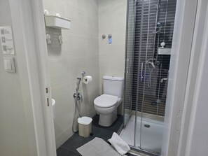A compact bathroom with a toilet, a shower enclosure, and a small trash bin. - Jualis Guest House (Porto)