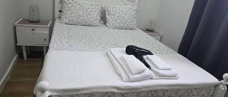 Cama bem arrumada com roupa de cama floral, mesa de cabeceira branca com abajur e cortina azul.