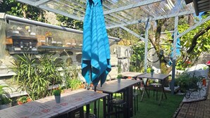 Ein Terrassenbereich im Freien mit Tischen und Stühlen, überdacht von einer blauen Markise, umgeben von Kübelpflanzen und einem kleinen Garten.