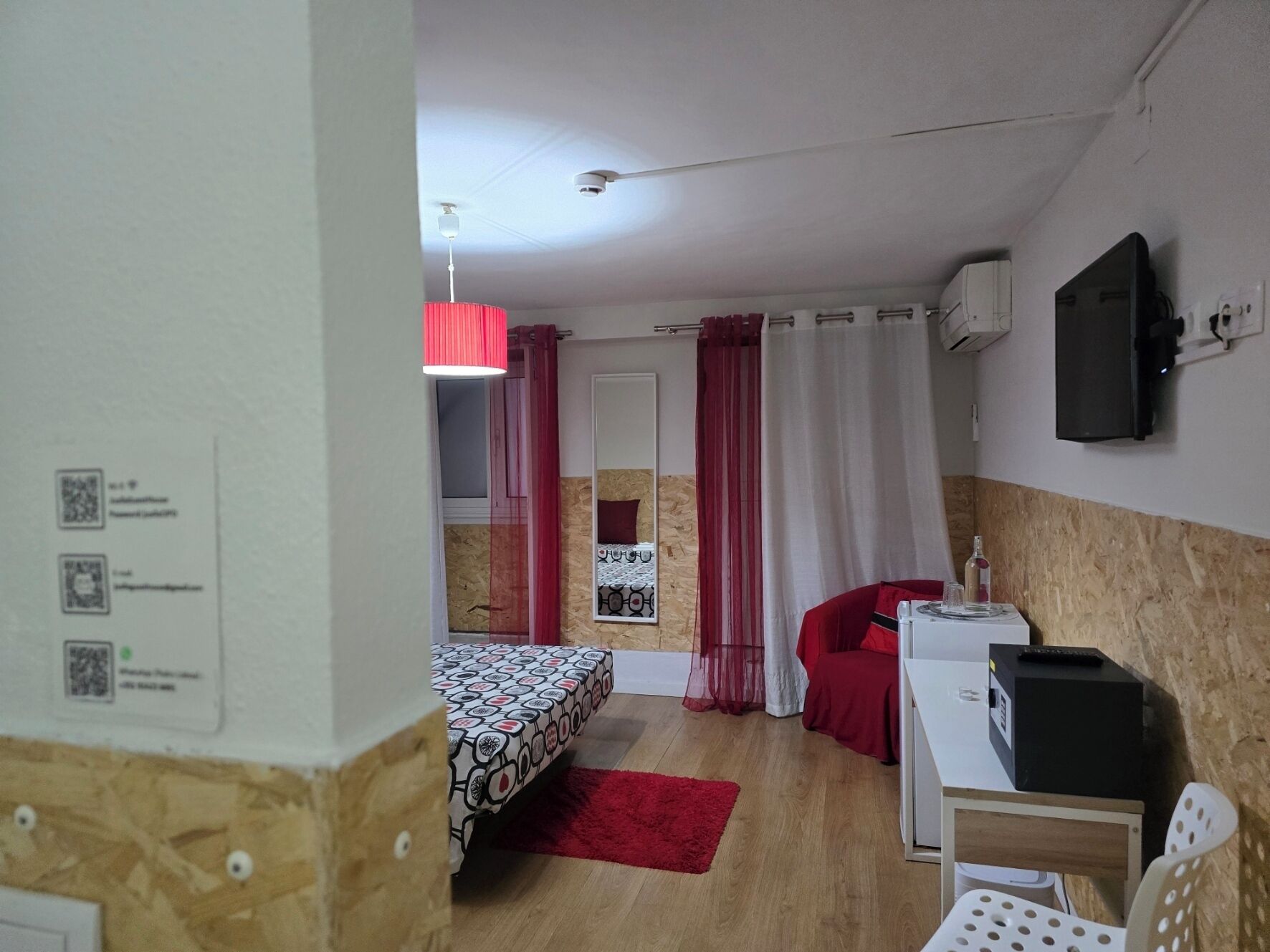 Ein kleines Zimmer mit Bett, Schreibtisch, Stuhl, Fernseher und einem roten Vorhang.