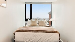Une chambre d’hôtel étroite avec une grande fenêtre, un lit recouvert d’une courtepointe blanche et marron, et une lampe sur la table de chevet.