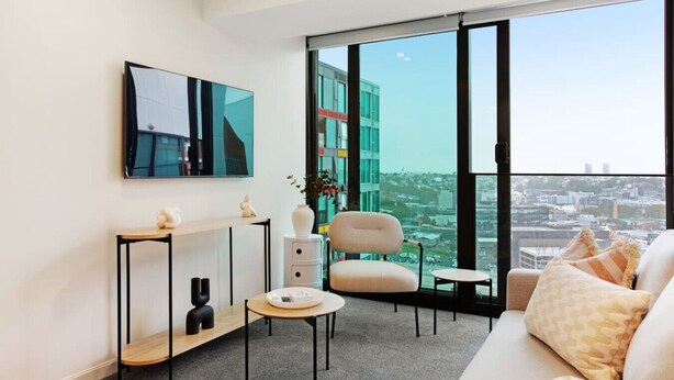 Un salon moderne comprenant un canapé, une table basse, un téléviseur fixé au mur et une grande fenêtre donnant sur la ville.