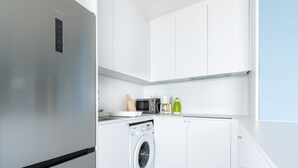 A modern kitchen with white cabinets, a stainless steel refrigerator, a washing machine, a microwave, and a coffee maker.