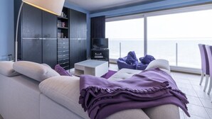 A modern living room with a sofa, a coffee table, and a TV. There is a bookshelf and a large window with a view of the sea.
