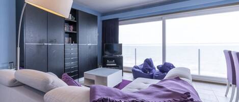 A modern living room with a sofa, a coffee table, and a TV. There is a bookshelf and a large window with a view of the sea.