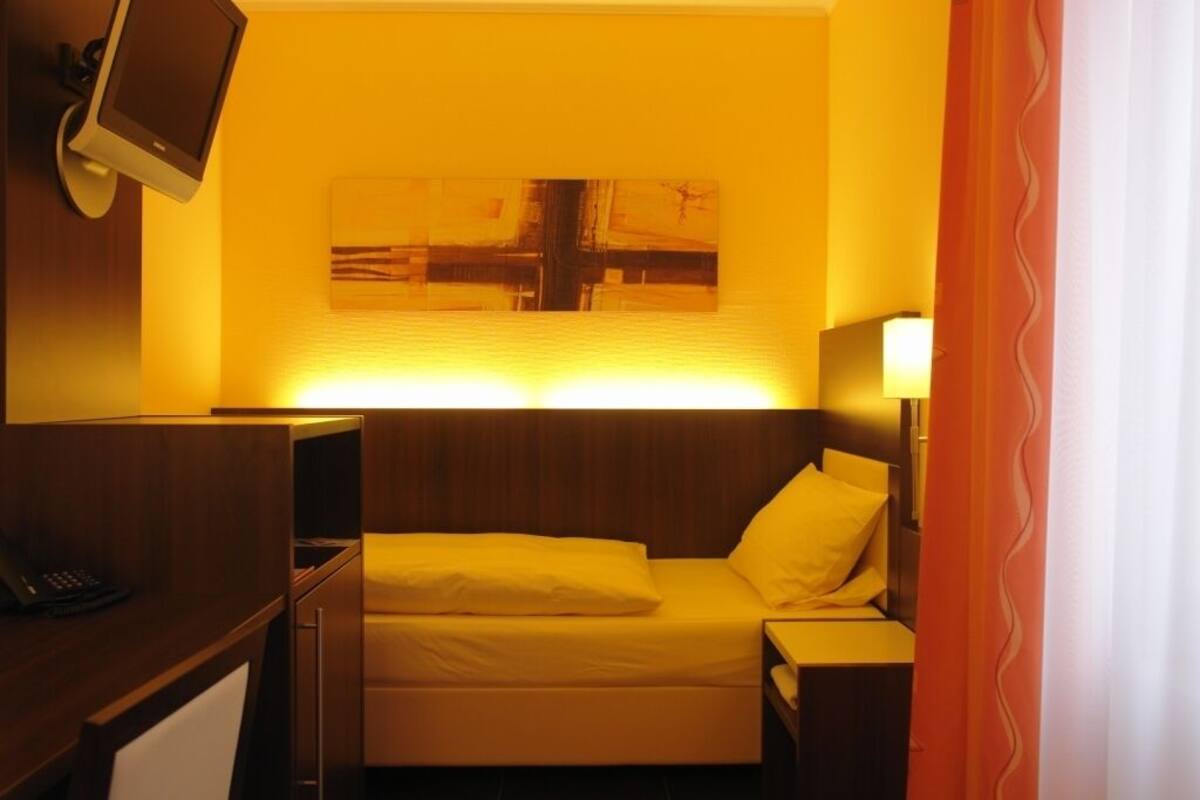 A hotel room with a bed, a desk with a computer, a TV mounted on the wall, and a painting above the bed.