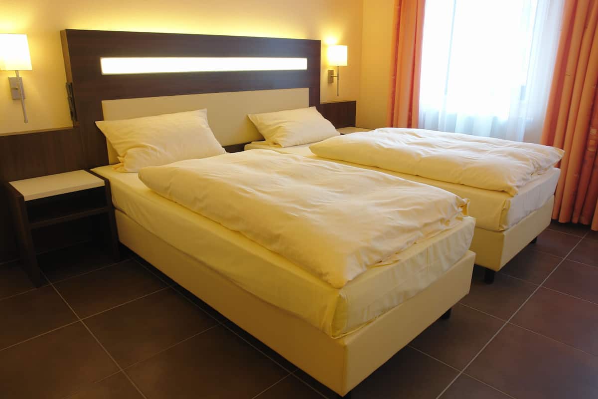 A hotel room with two beds, a nightstand, a wall-mounted lamp, and a window with curtains.