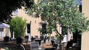 An outdoor patio area with tables and chairs, surrounded by olive trees and potted plants. - San Remo Fine.Food.Hotel (Nuremberg)