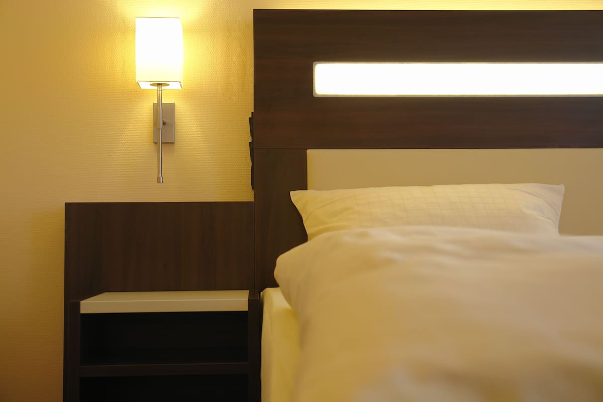 A hotel room with a neatly made bed, a bedside table, and a wall-mounted light fixture.