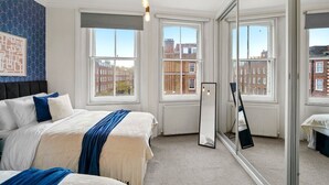 Quarto com uma cama grande, espelho e janelas amplas com vista para prédios.
