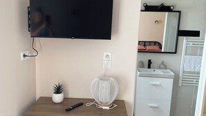 A bedroom with a TV mounted on the wall, a bedside table with a lamp, a small plant, and a wooden dresser.