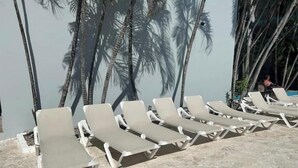 A row of beige sun loungers aligned on a stone patio with palm tree shadows cast on a light-colored wall. - Wind Chimes-Condo in Cabarete DR (Cabarete)