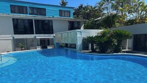 A swimming pool with a clear blue water surface, surrounded by a concrete deck. There are palm trees and a building with large windows in the background. - Wind Chimes-Condo in Cabarete DR (Cabarete)
