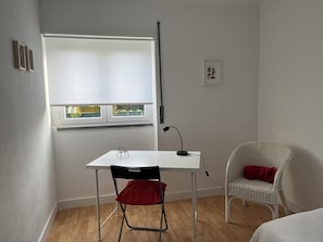 Una habitación pequeña y luminosa con un escritorio blanco, una silla de mimbre con un cojín rojo y una ventana con persianas.