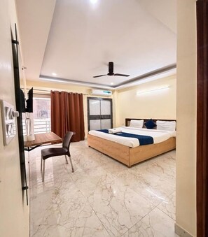 A hotel room with a bed, a desk, a chair, a ceiling fan, and a window with curtains. - Green Vista Stays (Gurugram)