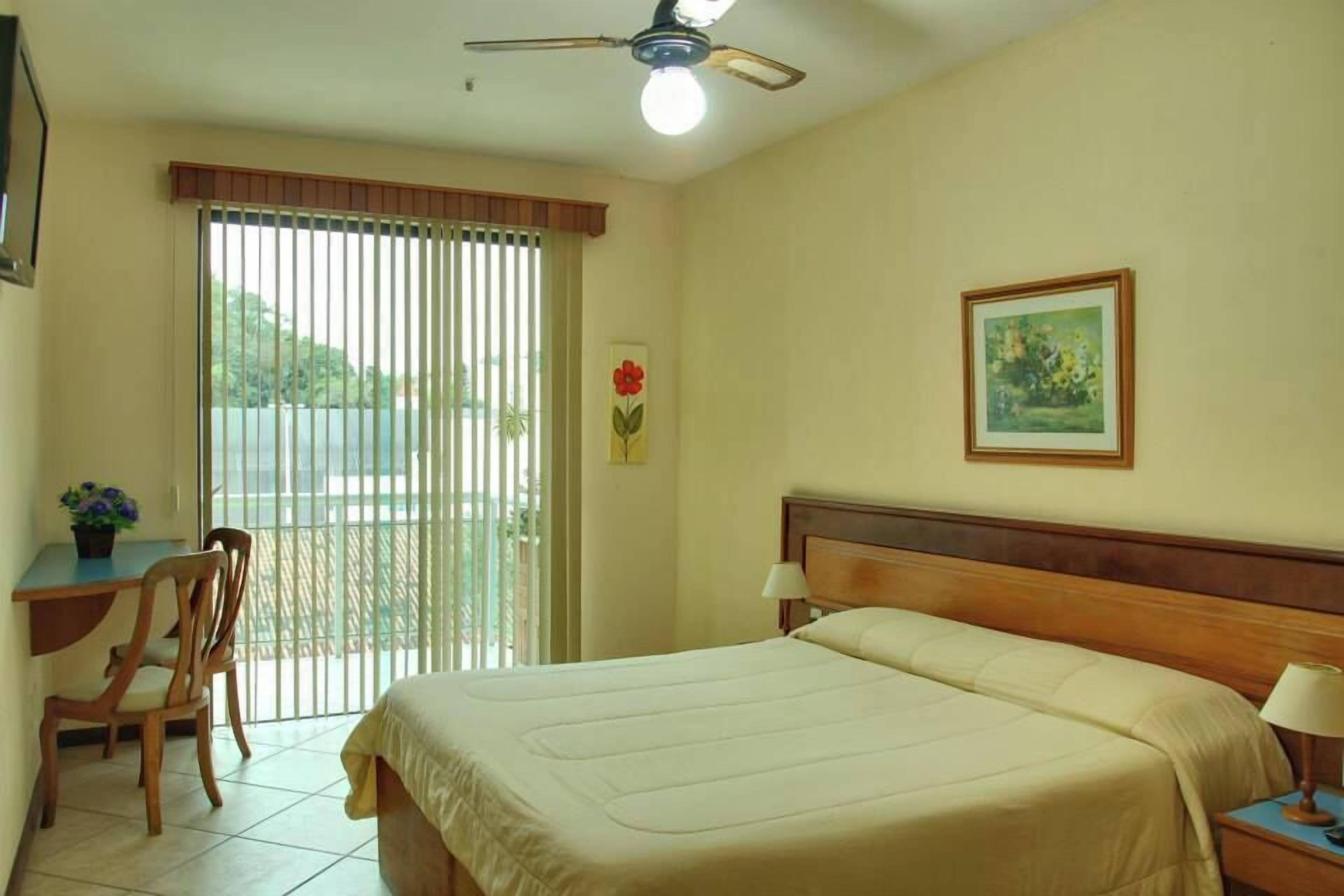 Habitación de hotel con cama, escritorio con silla, ventana con persianas, ventilador de techo y un cuadro en la pared.