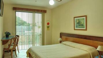 Habitación de hotel con cama, escritorio con silla, ventana con persianas, ventilador de techo y un cuadro en la pared.