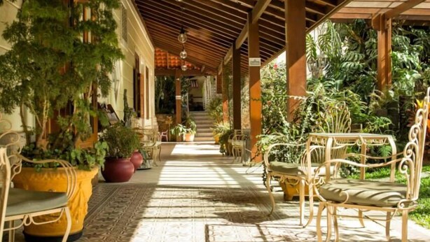 A covered outdoor area with a patterned floor, potted plants, and wicker furniture. - Hotel Dominguez Plaza (Nova Friburgo)