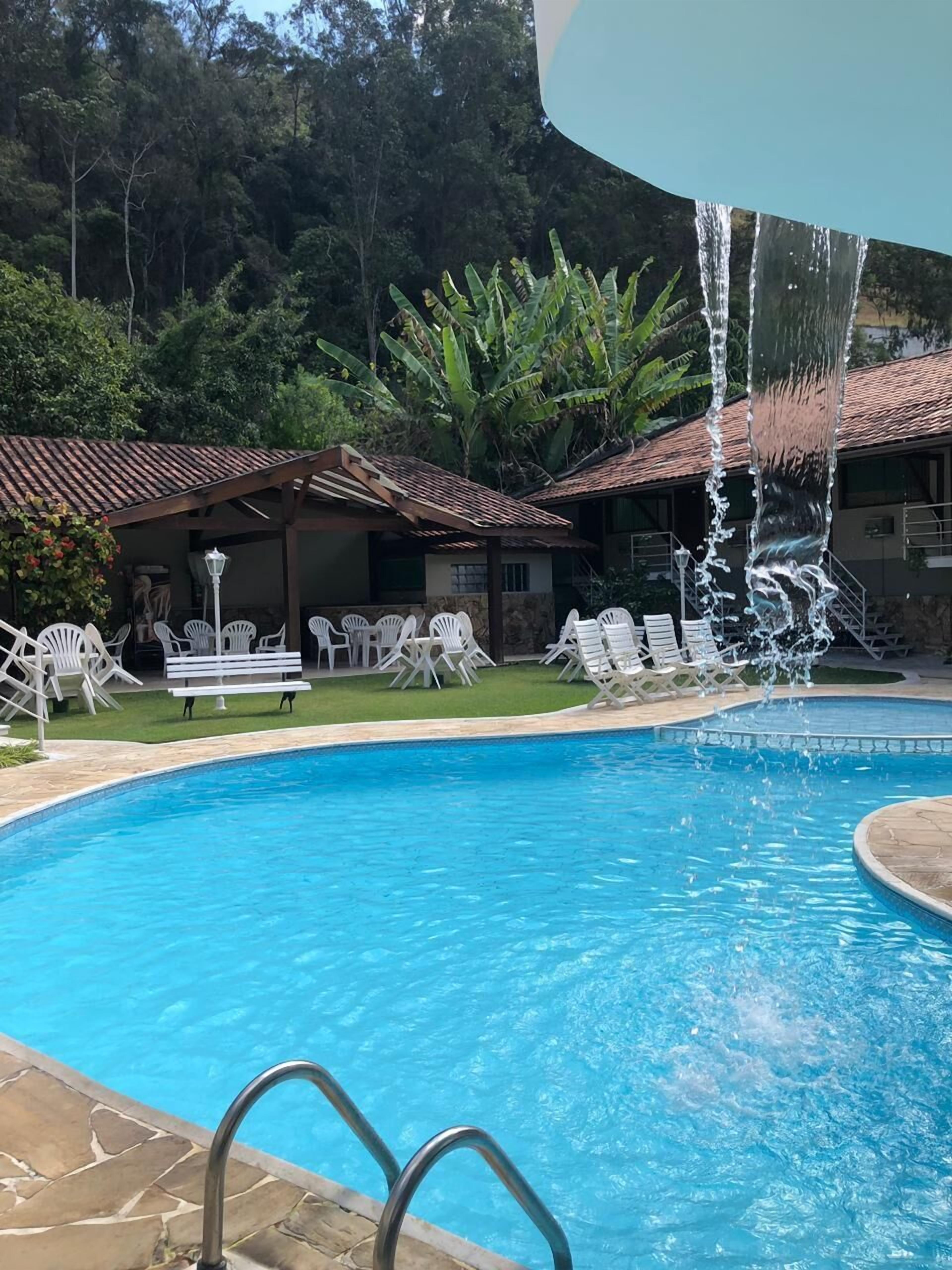 Piscina com fonte de água, rodeada por espreguiçadeiras brancas e uma área de estar coberta.